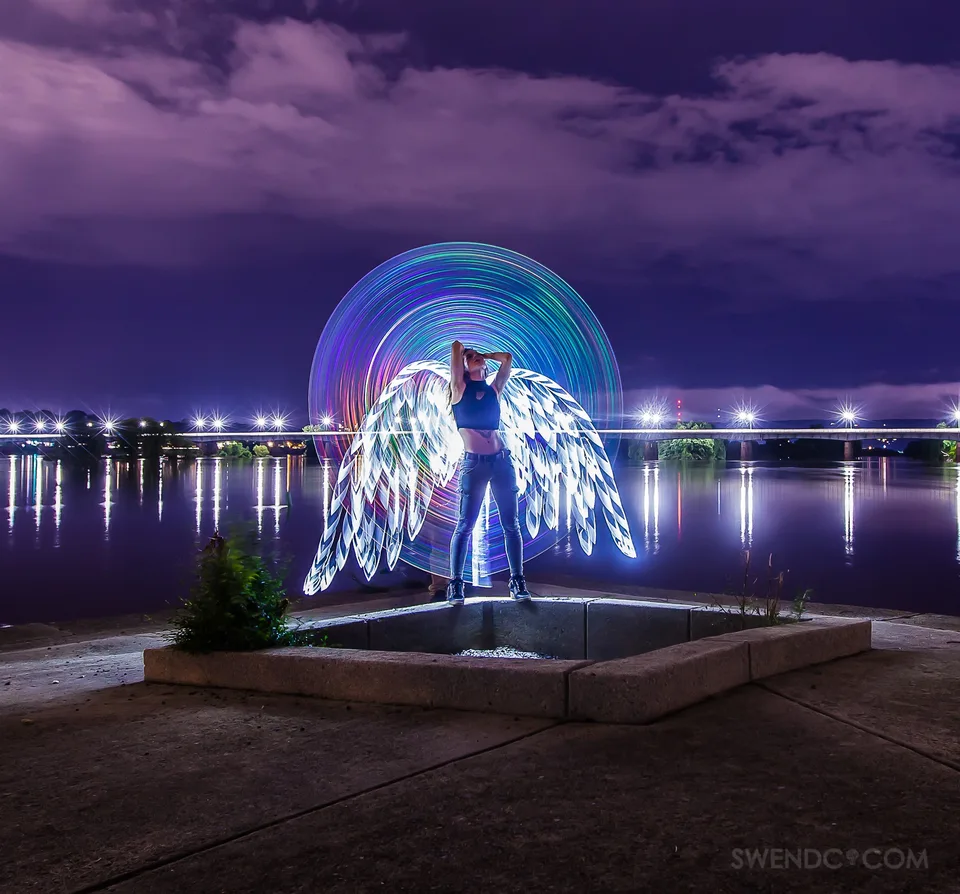 A woman stands inside a rainbow halo with bright light-painted wings beside the water at night.