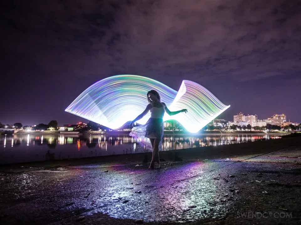 A woman in a white dress stands beneath a bright, flowing arc of rainbow light by the water.