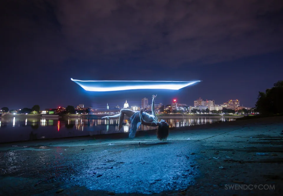 A dancer appears to float beneath a bright horizontal beam of light beside a city river at night.
