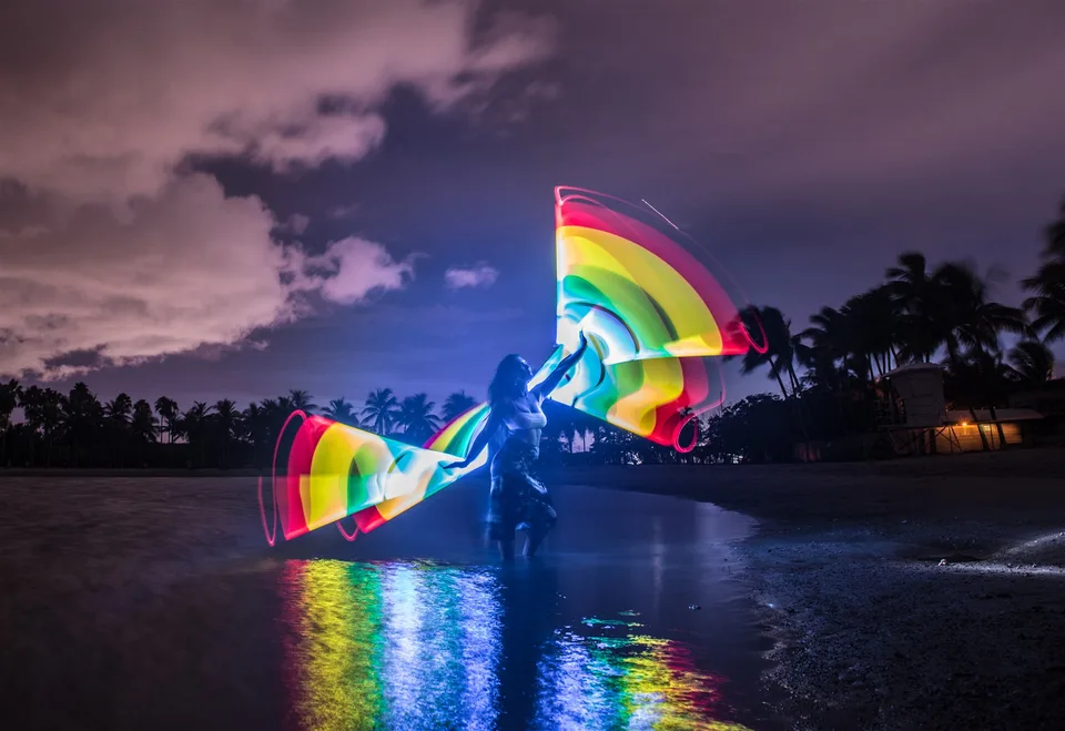 A performer paints a bright rainbow fan above shallow water, with color reflected on the surface.